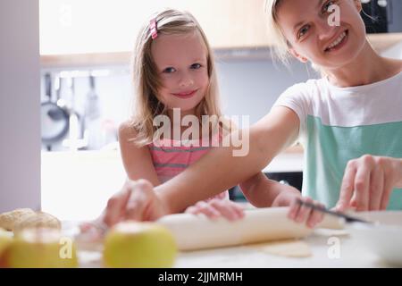 Glückliche junge Mutter und kleine Tochter rollt Teig auf Küchentisch mit Stift aus Stockfoto