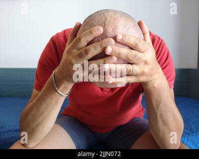 Eine Nahaufnahme eines verzweifelten kahlen Mannes weint und legt seine Hände auf seinen Kopf, der auf dem Bett sitzt Stockfoto