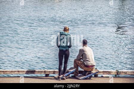 Paar auf dem Dock am Wasser mit einem Rad schweben Bretter - Mann mit Pullover und Mütze sitzen und Mädchen im Windbreaker stehen Stockfoto