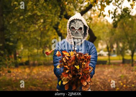 Unschärfe Halloween Menschen Porträt. Person in Sensenmann Maske auf Natur Herbst Hintergrund mit trockenen Blättern stehend. Halloween-Horror. Schädelgeist. Stockfoto