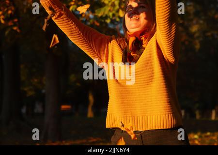 Unschärfe hallo Herbst. Fröhliche blonde elegante 40-jährige Frau draußen im Herbstpark, die den Herbst genießt und Blätter wirft. Viele fliegen orange, yello Stockfoto