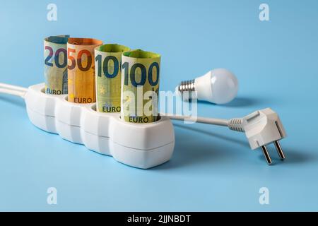 Anstieg der Energiepreise. Steigende Stromrechnungen. Euro-Banknoten werden an eine Steckdose, eine Glühbirne und einen Netzstecker auf blauem Hintergrund angeschlossen. Stockfoto