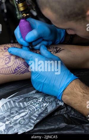 Eine vertikale Nahaufnahme der Hände eines Mannes, der in einem Tattoo-Studio das Bein einer Frau auf einer Bahre tätowiert Stockfoto