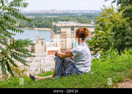 Eine junge Frau sitzt auf einem grasbewachsenen Feld auf einem Hügel und blickt unscharf in die Ferne über einen Fluss auf ein Stadtbild. Speicherplatz kopieren. Stockfoto