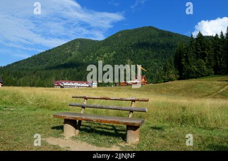 Eine hölzerne Bank auf einem ländlichen grünen Feld mit Bäumen in der Nähe von Bergen unter einem blauen Himmel Stockfoto