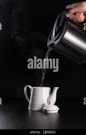 Eine vertikale Nahaufnahme der Hand, die das kochende Wasser in einen Krug auf schwarzem Hintergrund gießt. Stockfoto