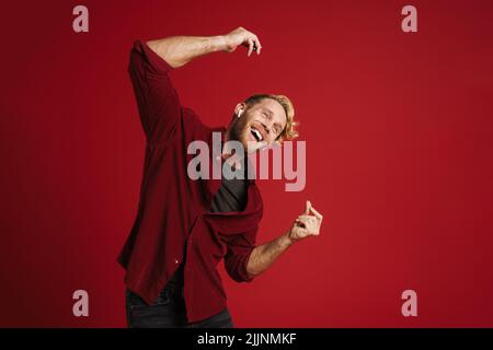 Blonde fröhliche Mann Musik hören und tanzen isoliert über rote Wand Stockfoto