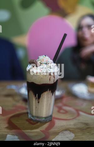 Eine Nahaufnahme einer Tasse Milchshake mit einem schwarzen Strohhalm auf dem Tisch mit einem lächelnden Mädchen im unscharfen Hintergrund Stockfoto