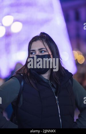 Eine flache Fokusaufnahme einer kaukasischen Frau, die eine schwarze Jacke mit schwarzen Ärmeln und eine schwarze Gesichtsmaske trägt, mit verschwommenen violetten Bokeh-Lichtern im Hintergrund Stockfoto