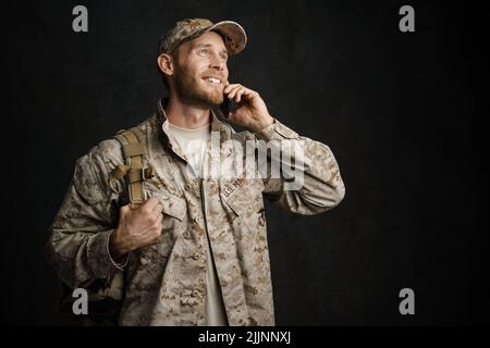 Weißer Militärangehöriger in Uniform lächelt und spricht auf dem Handy isoliert über der schwarzen Wand Stockfoto