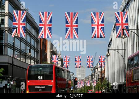 London, England - 2022. Juni: Über der Oxford Street hängen Reihen von Union Jack-Flaggen, rote Londoner Busse fahren entlang der Straße Stockfoto