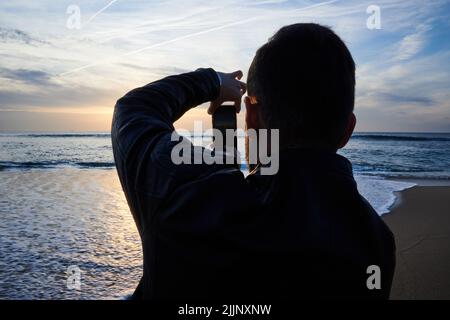 Rückansicht eines jungen Mannes, der den Horizont über dem Meer fotografiert Stockfoto