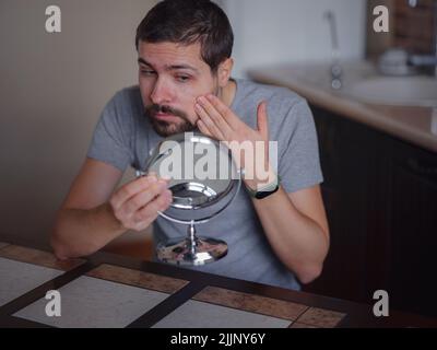 Kerl überprüft seine Haut. Der junge Mann, der im Spiegel schaut, berührt das Gesicht mit einem Pickel und fühlt sich verärgert. Anti-Akne Hautpflege Behandlung, hormonelles Ungleichgewicht, Stress, Ernährung, Hautproblem, Sommersprossen Aussehen Konzept Stockfoto