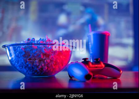 Joystick, Gamepad, Popcorn in einer großen Glasschale, Limonade in einem blauen Kunststoffglas vor dem Hintergrund eines Plasma-TV-Bildschirms. Neonbeleuchtung. Aufz Stockfoto