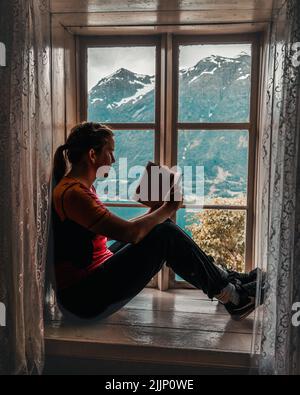 Vertikale Aufnahme eines Mädchens auf der Fensterbank beim Lesen eines Buches vor einer atemberaubenden Landschaft Stockfoto