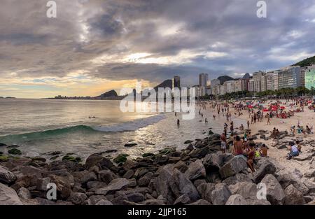 Die Welle kommt unter einem dramatischen, bewölkten Himmel mit schweren Wolken und Sonnenstrahlen, die die Menschen am Strand von der Küste von der Küste von der Küste beleuchten. Stockfoto