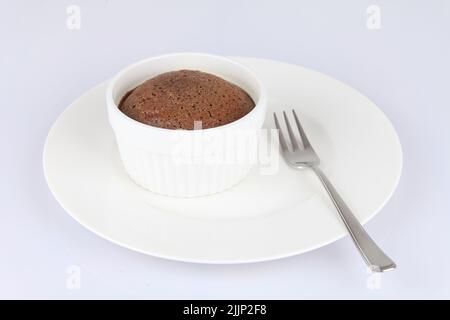 Ein Schokoladen-Souffle in einem klassischen weißen Porzellan-Ramekin auf einem weißen Teller mit einer Gabel Stockfoto