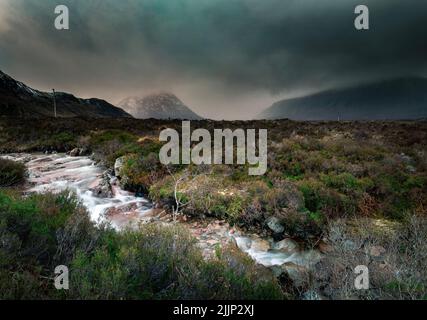 Eine malerische Aussicht auf einen Bach in einem Tal, umgeben von Bergen im Nebel mit dramatischen Wolken, Schottland Stockfoto