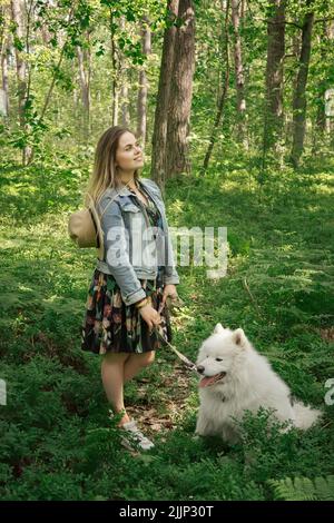 Junge schöne Frau, die im Wald aus der Stadt läuft, ihr weißer Hund samoyed. Stockfoto