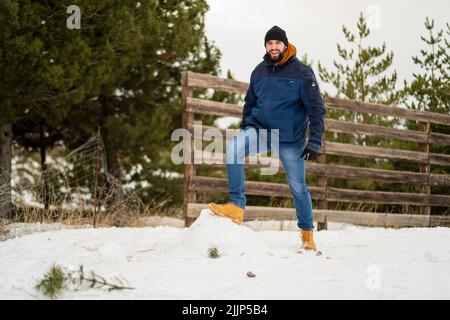 Ein junger Kaukasusmann, der im Winter auf dem Schnee im Park steht und lächelt Stockfoto
