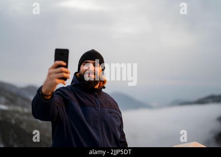 Ein junger Kaukasusmann in einer blauen Jacke lächelt und nimmt ein Selfie mit seinem Handy in der Natur Stockfoto