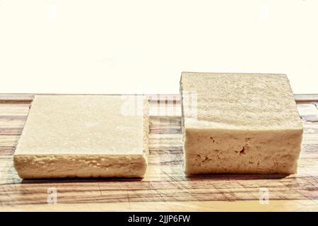 Zwei Tofu-Stücke - eines aus der Verpackung und das andere, das gedrückt wurde, um Wasser zu entfernen - sitzen auf einem Schneidebrett mit weißem Hintergrund - Platz für CO Stockfoto