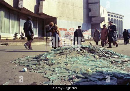 Bukarest, Rumänien, Januar 1989. Menschen, die an zerbrochenem Glas und Trümmern auf dem Universitätsplatz am Intercontinental Hotel vorbeikommen, Tage nach der antikommunistischen Revolution im Dezember 1989. Auf der Wand dahinter steht: 'Nieder mit dem Kommunismus' & 'Werft eure (Kommunistische Partei-)IDs weg'. Stockfoto