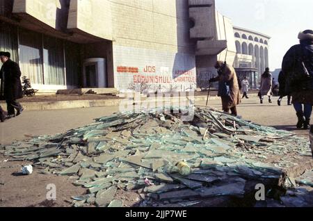 Bukarest, Rumänien, Januar 1989. Menschen, die an zerbrochenem Glas und Trümmern auf dem Universitätsplatz am Intercontinental Hotel vorbeikommen, Tage nach der antikommunistischen Revolution im Dezember 1989. Auf der Wand dahinter steht: 'Nieder mit dem Kommunismus' & 'Werft eure (Kommunistische Partei-)IDs weg'. Stockfoto