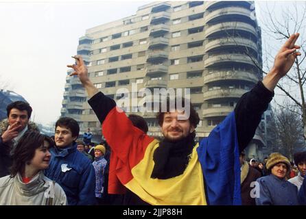 Bukarest, Rumänien, Januar 1990. Nach der antikommunistischen Revolution vom 1989. Dezember versammelten sich täglich Menschen auf dem Universitätsplatz, um gegen die neuen Menschen an der Macht zu protestieren, die die ehemaligen kommunistischen Beamten waren. Ein junger Mann macht das Siegeszeichen, trägt die Nationalflagge mit abgeschnittenem sozialistischem Emblem, ein Symbol während der Revolution, das das Ende der kommunistischen Ära darstellt. Stockfoto
