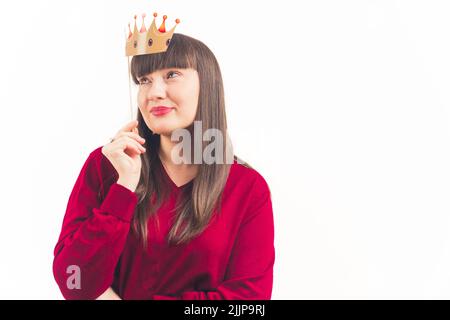 Weiße dunkelhaarige Frau mit Pony und weichem Make-up trägt roten Pullover mit gefälschter Papierkrone über ihrem Gesicht. Studioaufnahme. Isolierter Kopierbereich. Hochwertige Fotos Stockfoto
