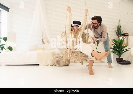 Junges weißes Paar im Schlafzimmer im Boho-Stil. Blonde Frau in VR-Brille auf hängendem Stuhl, gehalten von einem Mann mit dunklem Haarbart und Brille. Innenaufnahme. Hochwertige Fotos Stockfoto