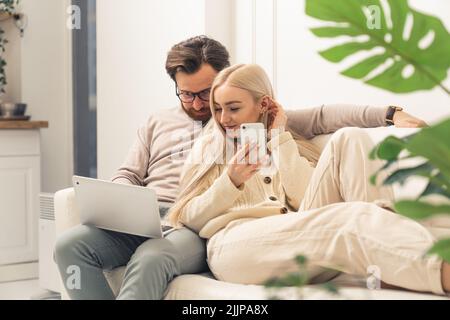 Kaukasisches Paar, das sich gemeinsam auf der Couch entspannt und einen Laptop auf dem Schoß des Mannes und die Frau mit dem Smartphone betrachtet. Innenaufnahme. Hochwertige Fotos Stockfoto