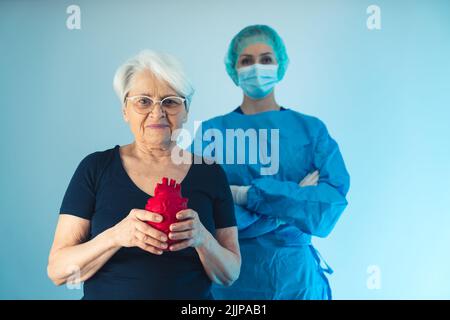 Alte kaukasische Frau, die vor einem Chirurgen-Arzt steht, während sie ein rotes künstliches Herz vor ihrer Brust hält. Hochwertige Fotos Stockfoto