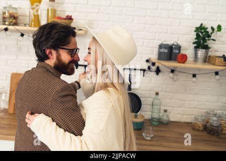 Dunkelhaariger weißer Mann mit Bart und Brille und blonde weiße Frau mit Hut tanzen langsam in der Küche lächelnd. Innenaufnahme. Hochwertige Fotos Stockfoto
