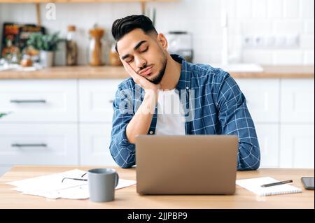 Müde überarbeiteter arabischer oder indischer junger Mann, Firmenangestellter, Student, der zuhause mit einem Laptop arbeitete, schlief während der Arbeit ein, müde von langweiligen Projekten, chronischem Schlafentzug, geschlossenen Augen Stockfoto