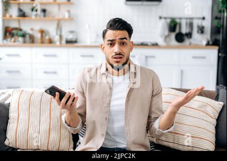 Verwirrte, verwirrte inder oder araber in legerer Kleidung, sitzt auf einem Sofa in einem Wohnzimmer, hält ein Smartphone in der Hand, blickt fragend auf die Kamera und spreizt seine Arme um sich Stockfoto