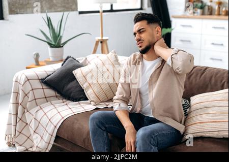 Konzept für Nackenschmerzen. Verstörter indischer oder arabischer Mann, der auf einem Sofa im Wohnzimmer sitzt, seinen Hals massiert und vor Schmerzen grimaziert, an Schmerzen leidet, braucht Ruhe und Behandlung Stockfoto