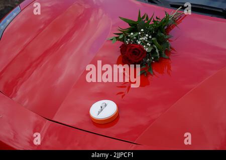 Ein Satz Verlobungsringe und ein Strauß roter Rosen auf einem roten Ferrari Stockfoto