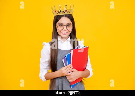 Schulmädchen in Schuluniform und Krone feiern den Sieg auf gelbem Hintergrund. Schulkind hält Bücher. Bildungsabschluss, Sieg und Erfolg Stockfoto