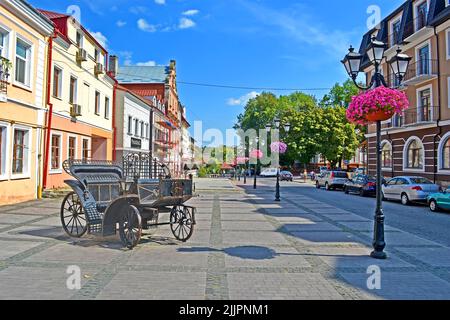 KAMENETS-PODOLSKY, UKRAINE - 25. AUG 2019: Stadtlandschaft Kamianets-Podilskyi mit geschmiedetem Wagen und Retro-Straßenlaterne mit Pylonschnur mit Petuni Stockfoto