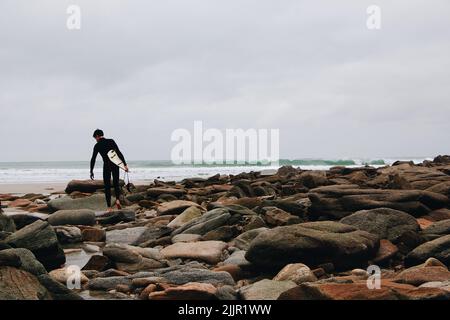 Ein junger Surfer, der über eine felsige Küste zum Meer läuft. Ein Bild, das mit VSCO mit einer voreinstellung von A6 verarbeitet wurde Stockfoto