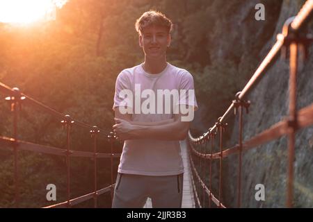 Ein flacher Fokus eines fröhlichen jungen, hübschen kaukasischen Mannes, der auf einer Brücke gegen die Sonnenstrahlen posiert Stockfoto