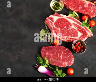 Verschiedene Steaks aus rohem Rindfleisch zum Grillieren mit Gewürzen und Geschirr auf dunklem Hintergrund, Draufsicht. Stockfoto