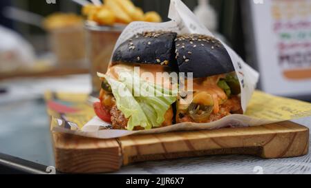 Eine Nahaufnahme eines leckeren schwarzen Brötchen-Burgers mit Fleisch, Salat, Tomaten und Sauce auf einem Tisch Stockfoto