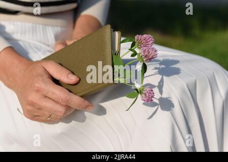 Beschnittene Ansicht einer verheirateten Frau in einem weißen Faltenrock, die ein geschlossenes olivfarbenes Tagebuch auf den Knien mit einem Blumenstrauß aus Kleeblatt im Inneren hält Stockfoto