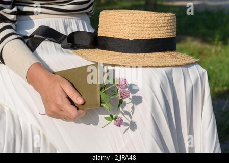 Verheiratete Frau, die in einem Park mit einem Bootsfahrer auf dem Schoß sitzt, mit geschlossenem olivfarbenem Tagebuch mit Blumenstrauß aus Kleeblatt im Inneren, beschnittene Ansicht. Stockfoto