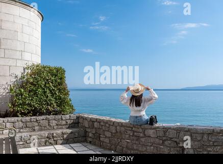 Eine junge Frau, die auf einer Steinmauer mit Blick auf das Meer sitzt Stockfoto