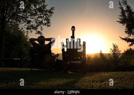 Rückansicht einer Frau, die während des Sonnenuntergangs in der Natur sitzt, mit Akustikgitarre neben ihr Stockfoto