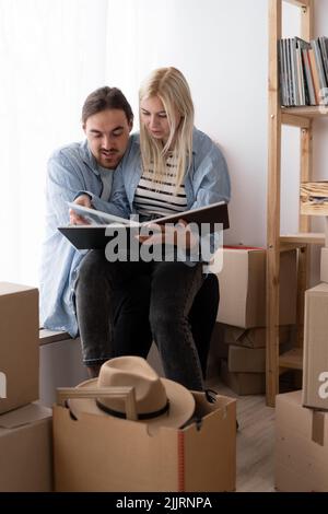 Nettes Paar, das Pappkartons in einem neuen Zuhause auspackend, auf dem Boden sitzend und ein Familienalbum angeschaut hat, bewegender Tag Stockfoto
