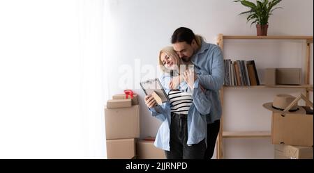 Junges Paar bewegen sich in neues Zuhause Stehen nehmen Auspacken Habseligkeiten aus Kartons hält Rahmen sieht Foto teilen Erinnerungen fühlt sich glücklich. Zuerst kaufen Stockfoto